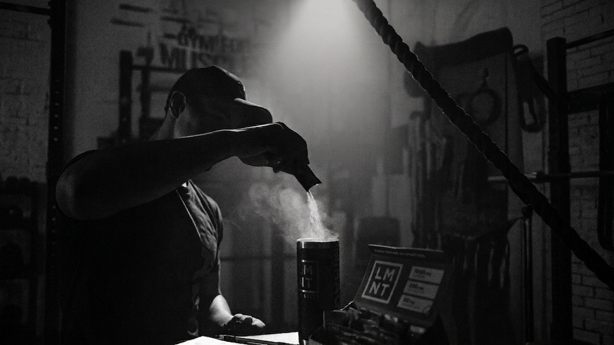 Man in a gym with a cap pouring powder into a shaker bottle, surrounded by workout equipment and dim lighting with a focused beam.