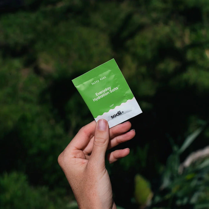 Hand holding Sodii's Salty Kiwi - 30 Sachets hydration salts pouch against a blurred natural green background