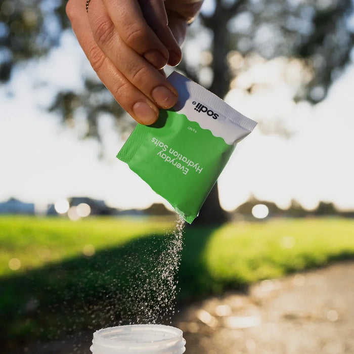 Hand pouring Salty Kiwi - 30 Sachets electrolyte hydration salt by Sodii into a bottle outdoors with greenery background