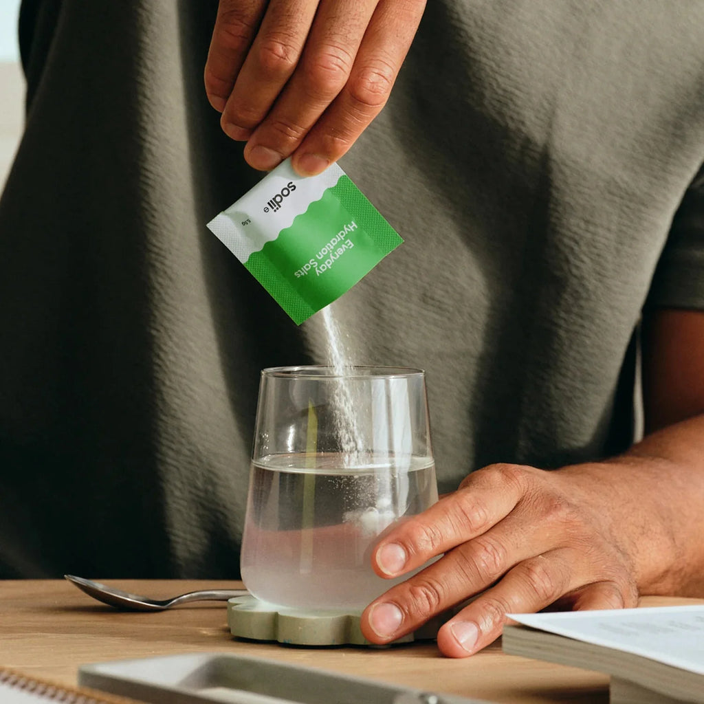 Hands pouring Salty Kiwi - 30 Sachets by Sodii electrolyte powder into glass water, on wooden table with book nearby