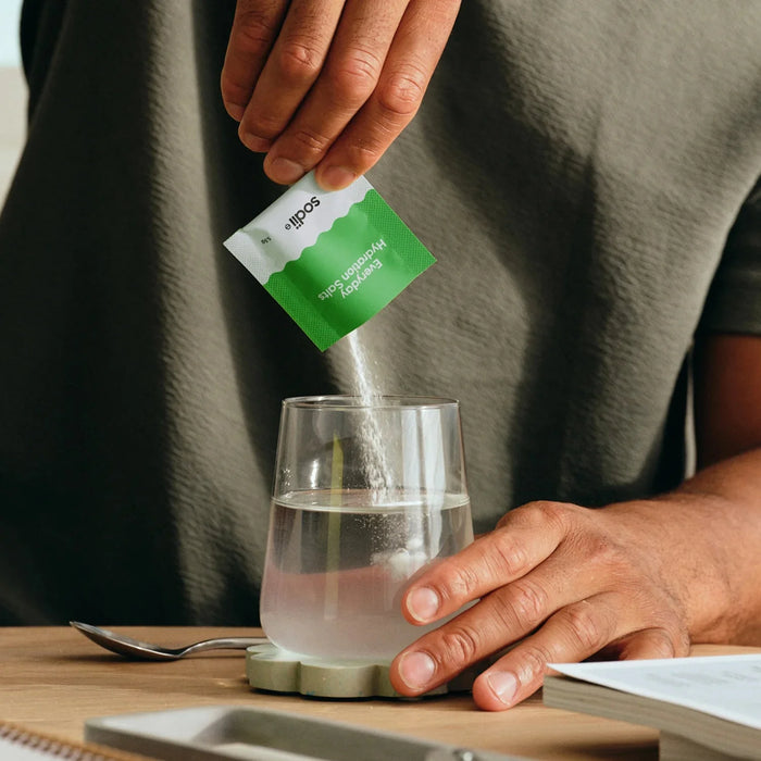 Hands pouring Salty Kiwi - 30 Sachets by Sodii electrolyte powder into glass water, on wooden table with book nearby