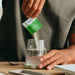Hands pouring Salty Kiwi - 30 Sachets by Sodii electrolyte powder into glass water, on wooden table with book nearby