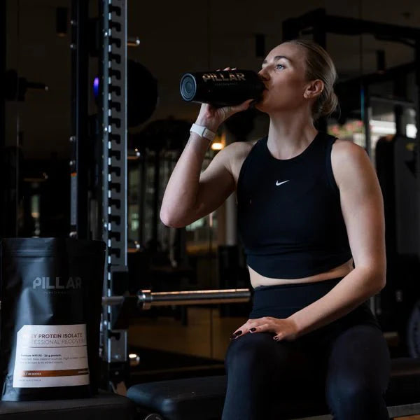 Woman in gym attire drinking from a shaker bottle with WPI - Vanilla - 1kg by PILLAR PERFORMANCE nearby